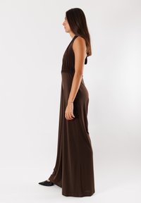 Abito maxi senza maniche marrone con vita elasticizzata, realizzato in un tessuto morbido. Il modello indossa scarpe nere con punta a punta. Design semplice e fluido.