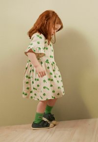 Beiges Kleid mit grünem Apfelmuster, Puffärmeln und gerafftem Rock. Grüne Strümpfe und schwarze Schuhe mit einer weißen Sohle. Holzboden als Hintergrund.