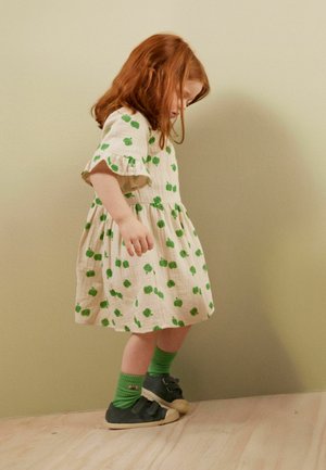 Beige mekko, jossa on vihreän omenan kuvio, puffihihat ja rypytetty hame. Vihreät sukkahousut ja mustat jalkineet, joissa on valkoinen pohja. Puulattia taustana.