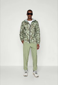 Jachetă ușoară cu model camouflage în nuanțe de verde, cu garnitură neagră; asortată cu pantaloni verzi și încălțăminte albă. Ochelari de soare purtați.