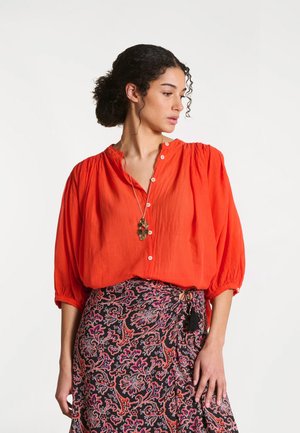 Femme aux cheveux bouclés portant un chemisier orange vif boutonné et une jupe portefeuille noire et rose à motifs, regardant sur le côté.