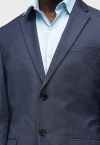 Άνδρας με σκούρο μπλε υφασμάτινο σακάκι πάνω από γαλάζιο πουκάμισο με κουμπιά, εστιασμένος στο στήθος και στην περιοχή του γιακά.