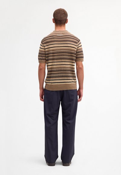 Uomo in piedi di spalle, indossa una maglia a maniche corte beige e marrone a righe lavorata a maglia con pantaloni larghi blu scuro.