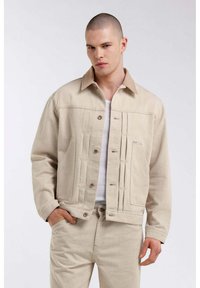 Giacca di jeans beige con colletto a contrasto, tasche frontali e chiusura con bottoni. Tessuto liscio con cuciture e dettagli ben visibili.