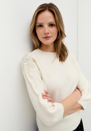 Cremefarbener Pullover mit Rundhalsausschnitt und dreiviertel Ärmeln. Mit floralen Perlenverzierungen auf der linken Schulter. Weiche Textur.