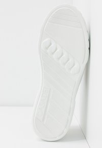 Podeszwa białego trampka z teksturowaną powierzchnią gumową, żłobionymi wzorami oraz wytłoczonym logo po boku. Gładki i elastyczny design.