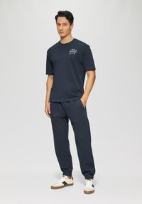 Navyfarbendes Baumwoll-T-Shirt mit kurzen Ärmeln und Logo, kombiniert mit passender Jogginghose. Die Sneaker haben ein weiß-schwarzes Design mit braunen Akzenten.