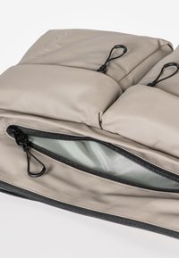 Borsa imbottita beige con due scomparti, dotata di una cerniera nera e linguette di estrazione. Tessuto liscio con una leggera lucentezza, fodera interna in una tonalità più chiara.