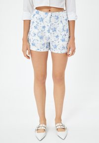 Bloemige shorts met blauwe patronen op een witte achtergrond, met een aansluitende taille en plooien, gecombineerd met witte puntige ballerina's.