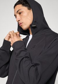 Μαύρο φούτερ με φερμουάρ, κατασκευασμένο από μαλακό ύφασμα, με κουκούλα με κορδόνι, φερμουάρ μπροστά και κεντημένο λογότυπο στο στήθος και το μανίκι.