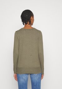 Olivengrüner Pullover mit langen Ärmeln, gerippten Bündchen und Saum. Glatte Textur, runder Ausschnitt und taillierte Passform. Getragen mit blauen Jeans.