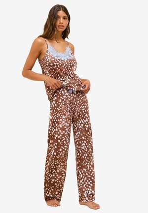 Frau trägt braun-weiß gepunktetes Pyjama-Set mit blauer Spitzenverzierung am ärmellosen Top, steht barfuß.
