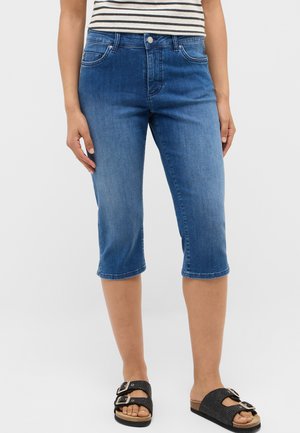 Woman wearing mid-blue cropped denim jeans, a striped top, and black double-buckle sandals against a plain background.