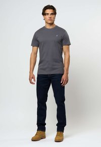 Harmaa lyhythihainen t-paita pyöreällä pääntiellä ja sinisellä brodeeratulla logolla, yhdistettynä tummansinisiin farkkuihin ja vaaleisiin nauhakenkiin.