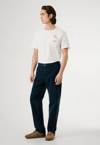 Wit katoenen T-shirt met een zakje en een grafische print, gecombineerd met marineblauwe corduroy broek. Schoeisel is bruin met subtiele accenten.