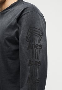 Κοντινό πλάνο ενός μακρυμάνικου σκούρου γκρι πουκαμίσου με κατακόρυφη γραφική στήλη και επαναλαμβανόμενο κείμενο "Aries" στο μανίκι.