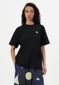 Camiseta de algodón negra de manga corta, con un pequeño emblema de flor blanca en el lado izquierdo del pecho. La tela tiene una textura suave.