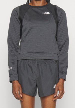 Person iført mørkegrå langærmet North Face-trøje og matchende shorts, stående mod en ensfarvet lys baggrund.