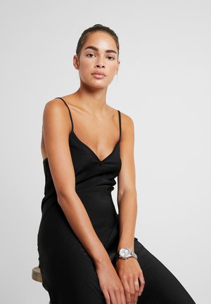 Vestido negro de tirantes finos, escote en V, tejido texturizado. Reloj de pulsera plateado con esfera redonda y correa de metal en la muñeca izquierda.