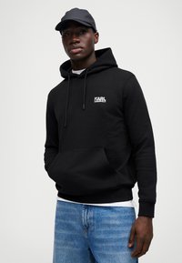 Μαύρη φούτερ με κουκούλα και μπροστινή τσέπη, με λευκό λογότυπο "KARL LAGERFELD". Συνοδεύεται από μαύρο καπέλο. Φοριέται με μπλε τζιν.