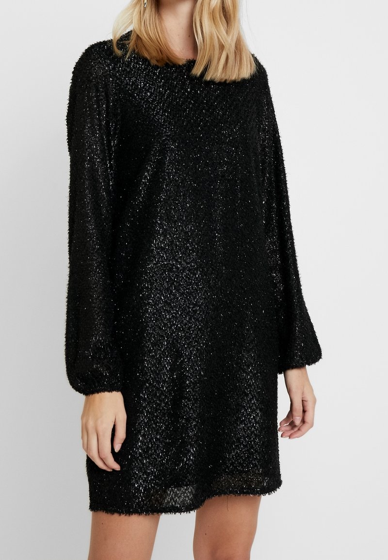 Femme portant une robe noire texturée scintillante à manches longues avec un col rond sur un fond clair uni.