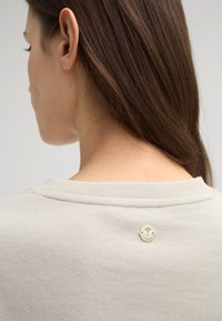 Light beige sweatshirt featuring a round, gold-tone logo accent on the back, with a soft texture and ribbed neckline.