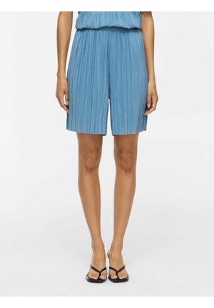 Femme portant un short plissé bleu et des sandales noires, debout devant un fond blanc uni.