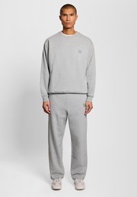 Mann steht frontal zur Kamera, trägt ein hellgraues Sweatshirt und passende Sweatpants sowie beige-weiße Sneaker, vor einfachem grauem Hintergrund.