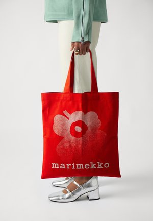 Röd canvas totebag med ett stort silverblomtryck och ordet "marimekko" i vitt. Väskan hålls av en person som bär silverskor.