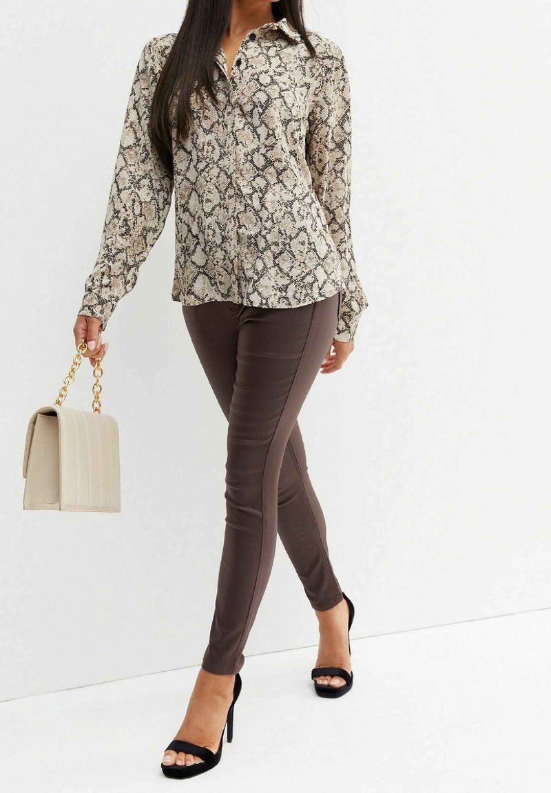 Skjorta med ormskinnsmönster och långa ärmar, kombinerad med åtsittande bruna byxor och svarta stiletto-klackar. Beiga handväskan har en guldkedjesnodd.