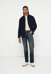 Camisa de botones de pana azul marino, camiseta beige debajo, jeans azules, zapatillas blancas con suelas beige, pose de pie, textura suave.
