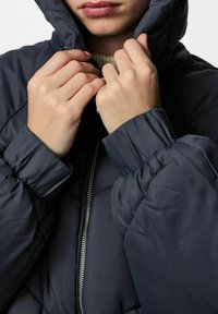 Tmavě modrá péřová bunda s kapucí, s prošívanou texturou, elastickými manžetami a předním zipem. Ruce upravují límec.