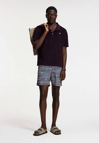 Tricou polo violet închis, pantaloni scurți de baie cu dungi albastre și albe, purtând o geantă bej, încălțat cu șlapi olive, stând în picioare pe un fundal deschis.