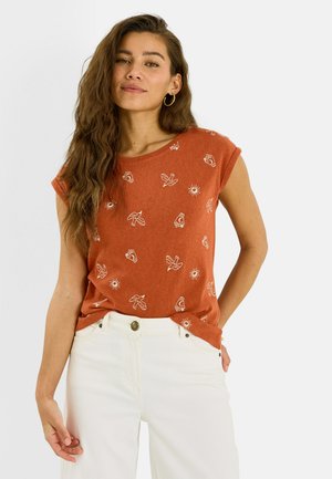 Femme aux longs cheveux ondulés portant une chemise orange rouille avec des imprimés blancs de soleil, d'oiseau et de main, associée à un pantalon blanc taille haute.