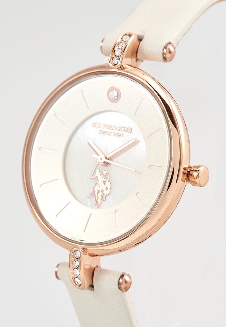 Reloj de oro rosa con correa de cuero blanca. Cuenta con una esfera de nácar, un logo de jugador de polo y acentos de cristal transparente en el bisel.