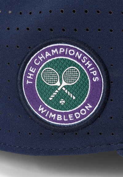 Temno modra kapa z perforirano tkanino, s krožno vvezano značko. Značka vključuje vzorec zelenih diamantov, vijolični rob in prekrižane loparje.