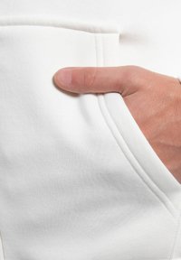 Un tessuto bianco di una felpa con cappuccio presenta una tasca frontale con una mano appoggiata, mettendo in evidenza la texture e i dettagli della cucitura.