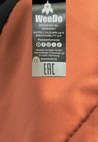 WeeDo-märke på orange texturerat tyg. Har vattensäule 10K, andningsförmåga 5K och märkesdetaljer med skötselråd.