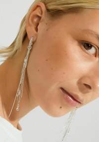 Blonde vrouw met lange, zilveren kettingoorbellen met kleine decoratieve elementen, die huidtextuur en natuurlijke make-up laat zien.