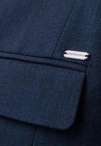 Tissu chevron bleu foncé avec une poche à rabat et une petite étiquette métallique argentée portant la mention "QUINTESSENCE" attachée au-dessus de la poche.