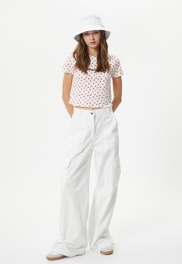 Sombrero blanco tipo bucket, camiseta corta de lunares rojos y blancos, y pantalones cargo blancos sueltos con bolsillos. Calzado casual con suela marrón.