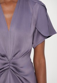 Blouse lavande avec un décolleté en V profond, des manches courtes et un détail de nœud froncé à la taille. Tissu lisse et texturé avec une légère brillance.