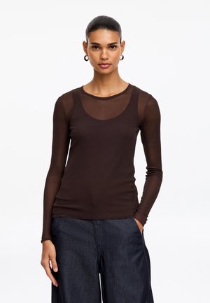 Mujer con el pelo oscuro recogido en un moño, que lleva una blusa negra transparente de manga larga sobre una camiseta negra y pantalones anchos de denim oscuro, mirando hacia adelante.