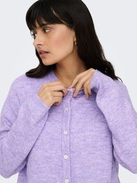 Lilla cardigan med knapper laget av mykt strikkestoff, med rund hals og ribbestrikkede mansjetter. Knapper er lyse og runde.