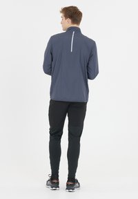 Veske som er vannavstøtende, med glidelås i mørkegrå farge og reflekterende bakstripe; kombinert med svarte smale bukser og grå løpesko.