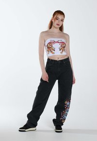 Top corto blanco sin mangas con gráficos de tigre, acompañado de unos jeans negros holgados que presentan bordados coloridos de calaveras y flores en una pierna.