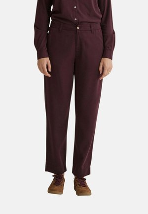 Persona que lleva pantalones morados oscuros de pierna recta, camisa morada oscura a juego con botones y zapatillas marrones con cordones morados.
