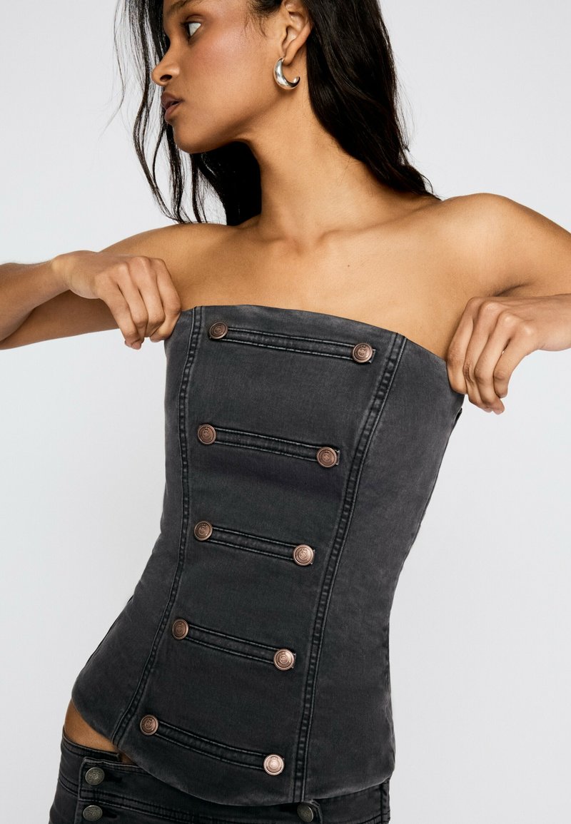 Femme ajustant un corset en denim foncé sans bretelles avec des boutons décoratifs, portant des boucles d'oreilles argentées en forme de créole, et regardant sur le côté.