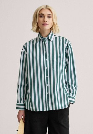 Lyshåret person iført grøn og hvid skjorte med lodrette striber og sorte bukser, der holder en stråhat i venstre hånd.