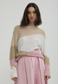 Donna con capelli scuri di lunghezza media che indossa un maglione color block beige, bianco e rosa e una gonna plissettata rosa su uno sfondo semplice.